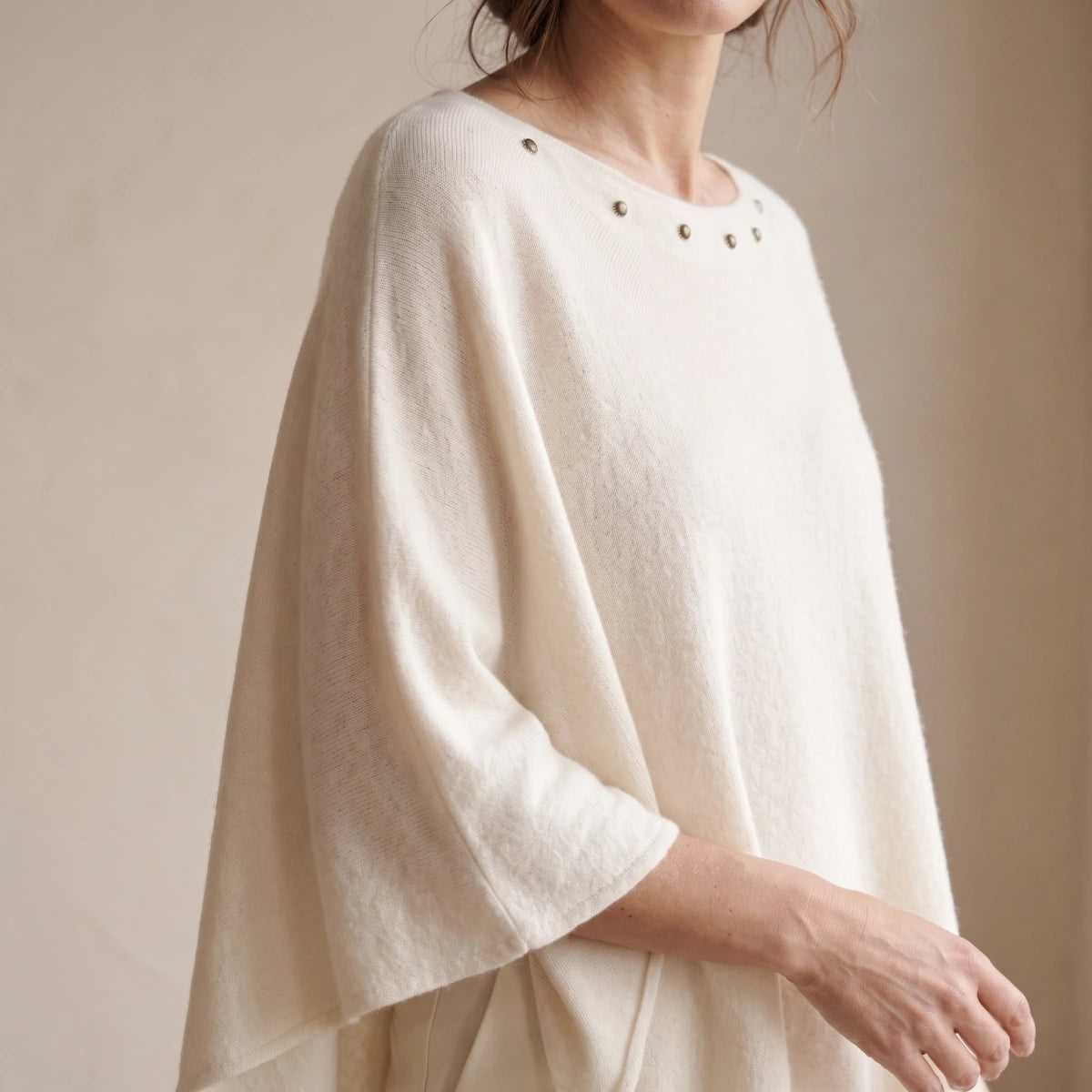 detalle de una mujer con poncho blanco de punto, escote ancho con tachuelas metálicas y manga amplia 