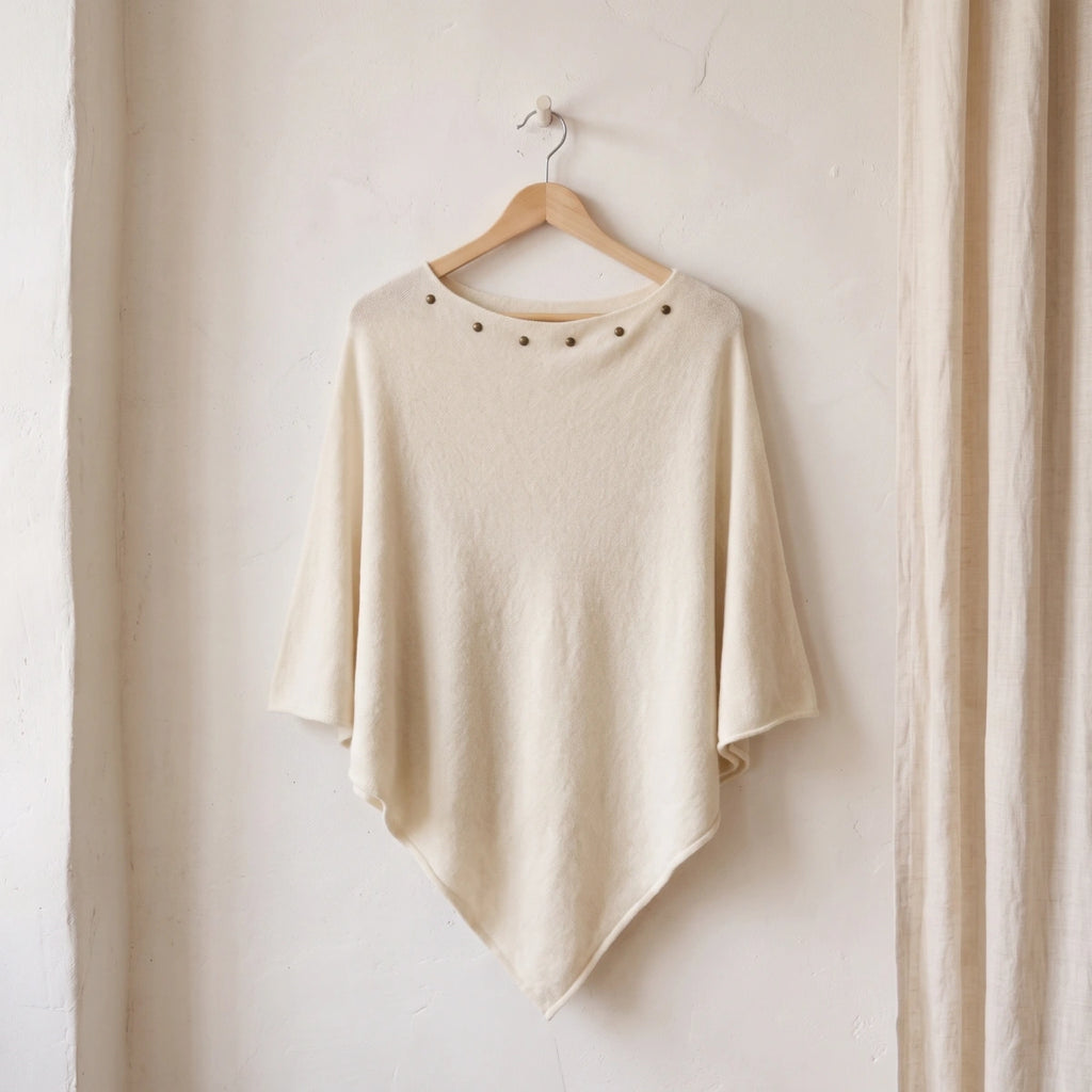 poncho blanco con tachuelas en el escote colgado de una percha de madera sobre una pared blanca Kloreys
