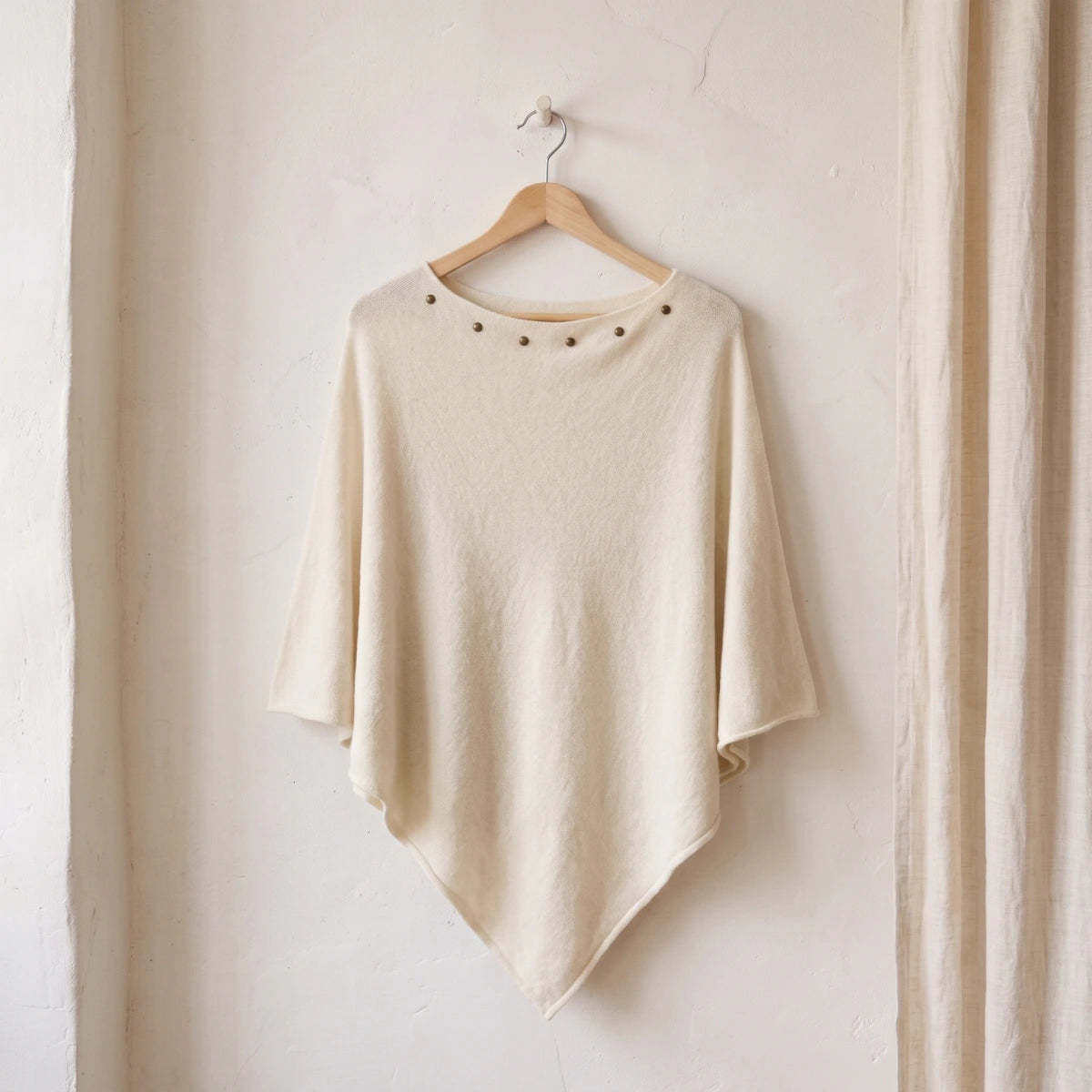 poncho blanco con tachuelas en el escote colgado de una percha de madera sobre una pared blanca Kloreys