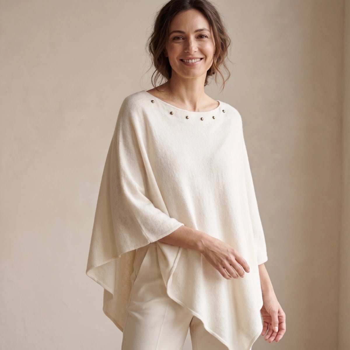 mujer sonriente con poncho blanco de punto con tachuelas y pantalón claro sobre fondo beige 