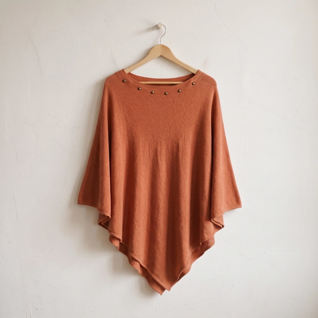 Poncho terracota con tachuelas colgado de una percha de madera sobre pared blanca Kloreys