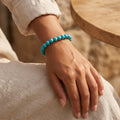 Pulsera elástica de howlita azul turquesa 8 mm en muñeca de mujer con ropa de lino